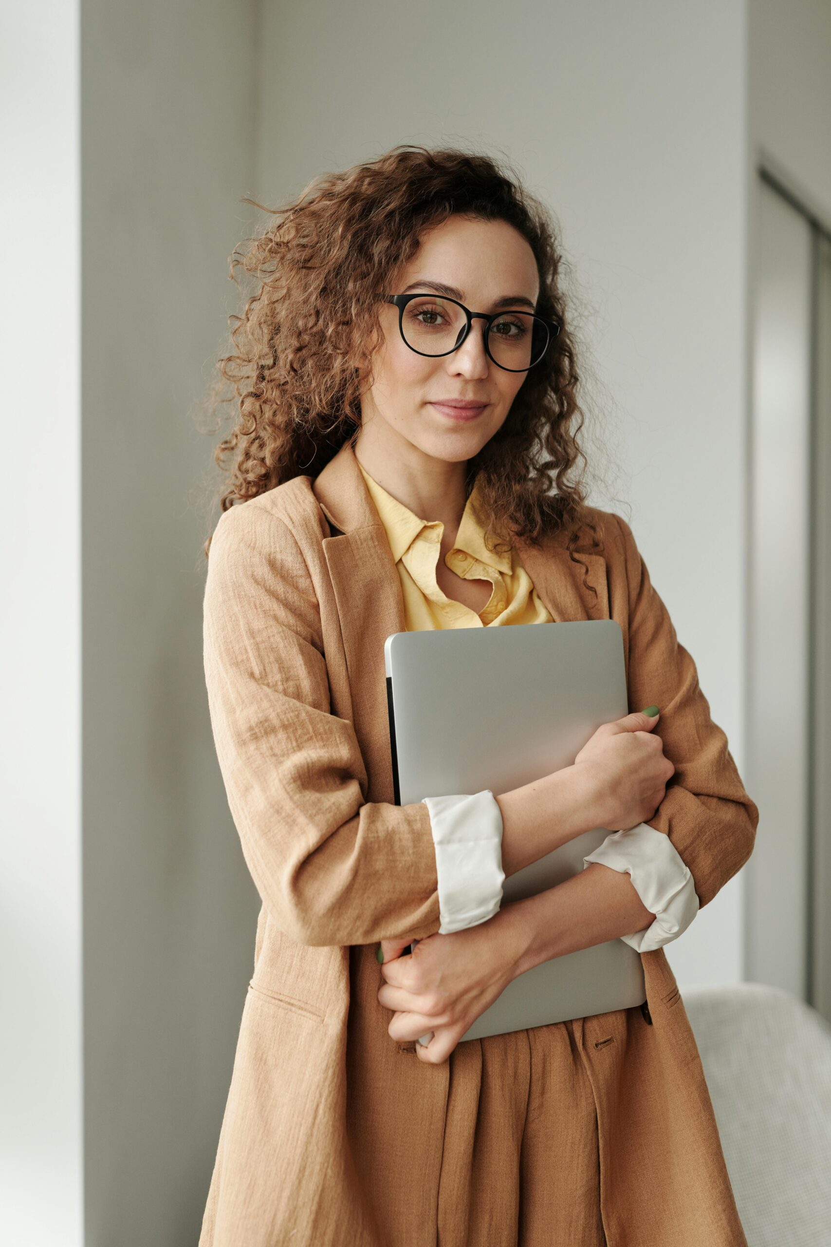 Our Team A confident woman in formal attire holding a laptop, standing in an office environment and looking at the camera.