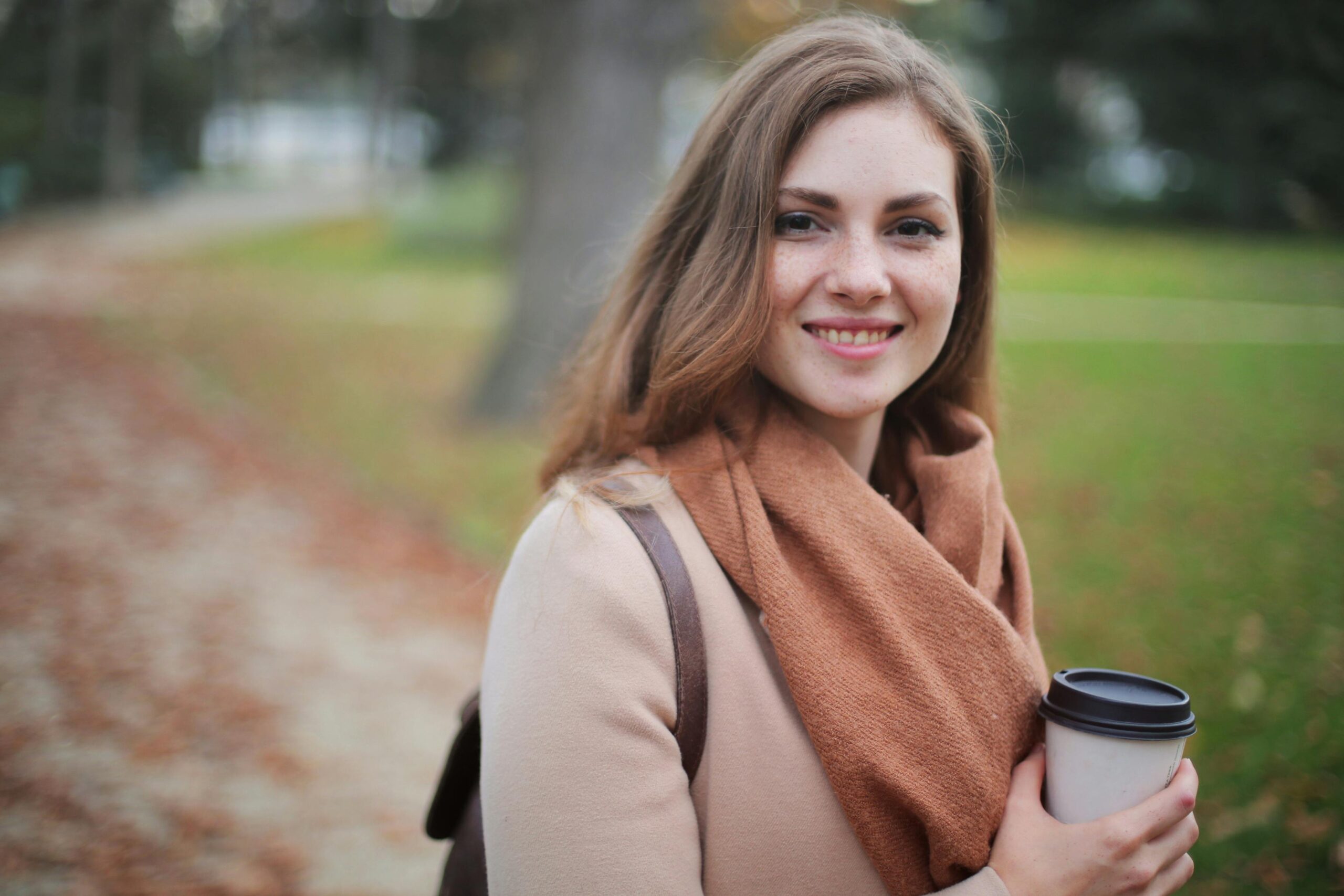 Our Team Portrait of a smiling woman holding a coffee cup in a serene autumn park setting.
