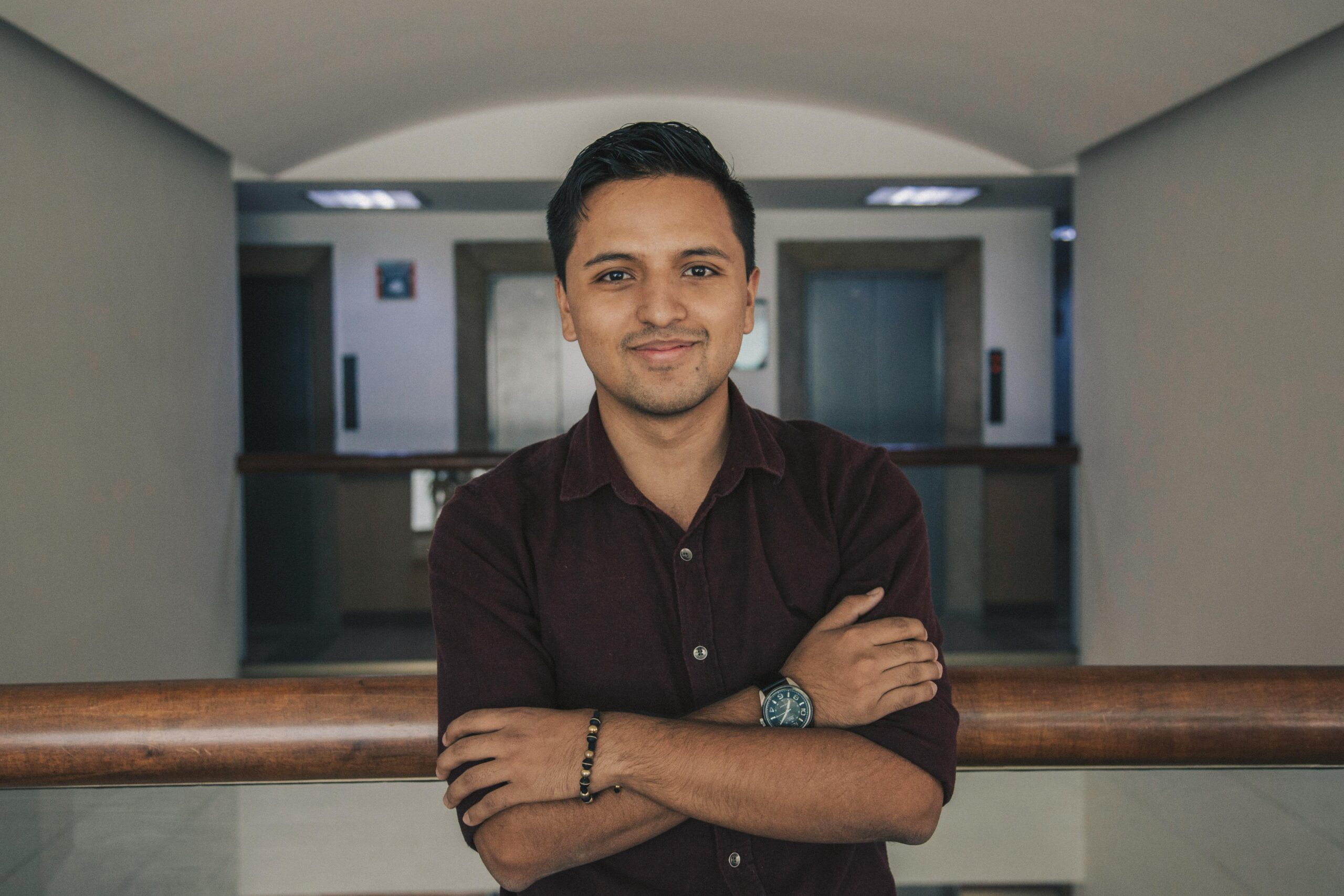 Our Team Positive Indian in casual outfit standing with crossed arms near glass fence while smiling and looking at camera in modern corridor